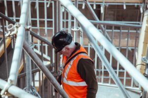 Construction worker in black hard hat, orange high-vis stood on scaffolding