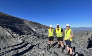 Three males in high vis jackets and hardhats stand on slate quarry grounds