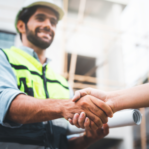 A male contractor with facial hear wears a high vis jacket and hardhat. His hand is outstretched shaking another hand