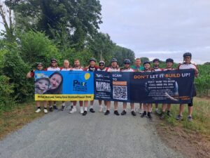 A group of cyclists stand on a road holding two banners promoting a charity