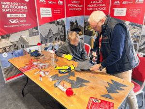 Small exhibition stand with young man sat at wooden desk being shown how to shape a slate roof tile by man stood up.
