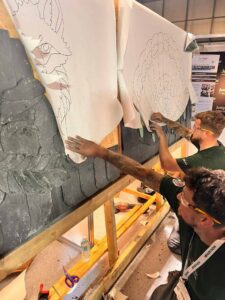 Great British Slate Off 2026 at the Installer Show finals with paper templates overlaying slates which have are being positioned and shaped to form fox and peacock designs.